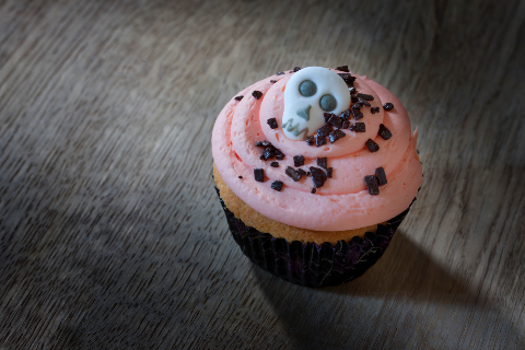 Pink frosted vanilla cupcake with black sprinkles and a sugar skull decoration on top. The cupcake is sitting on a grey wooden background.